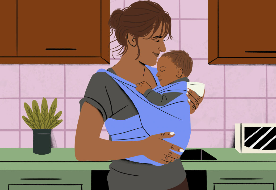 Smiling woman holding a sleeping baby in a blue sling in a cozy kitchen setting