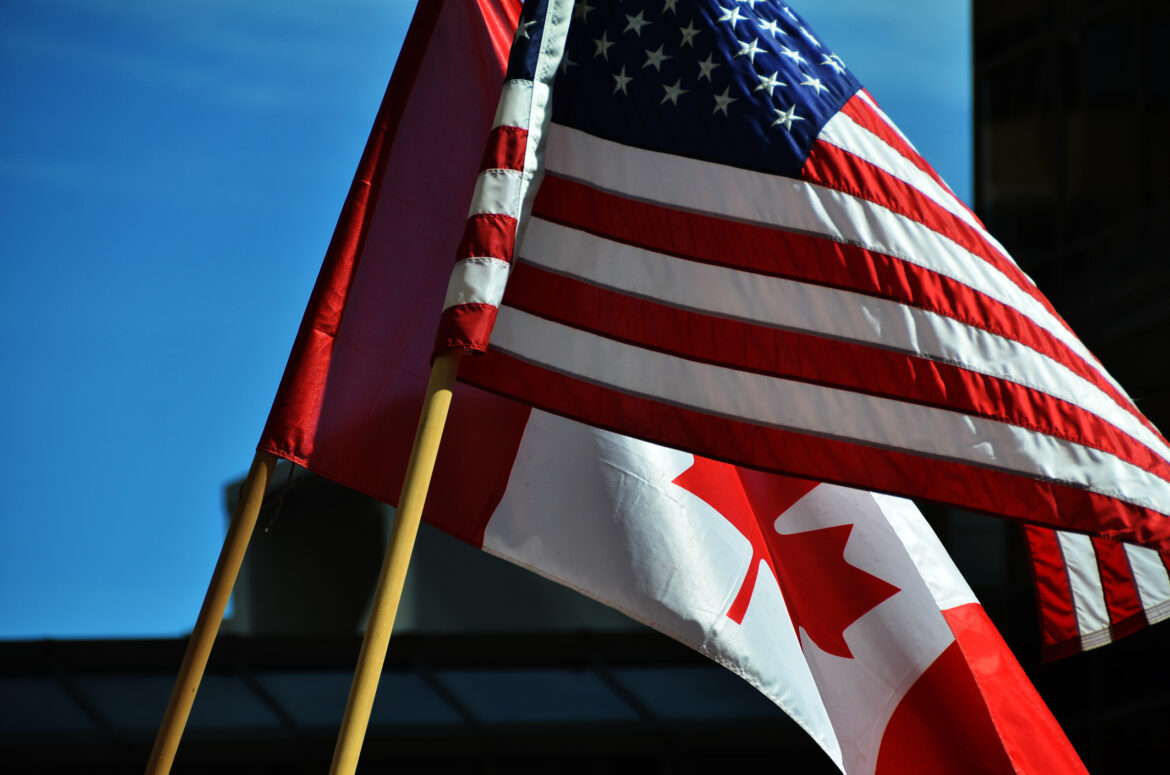 A close up of an American flag on a wooden pole waving in the wind directly in front of a Canadian flag on a wooden pole in an urban environment.